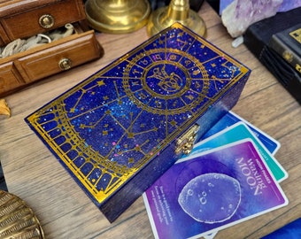 Puede incluir: Una caja de madera azul con una tapa dorada que presenta un diseño celestial con constelaciones y símbolos del zodíaco. La caja está abierta y revela un juego de cartas de tarot con un diseño de acuarela púrpura y azul.