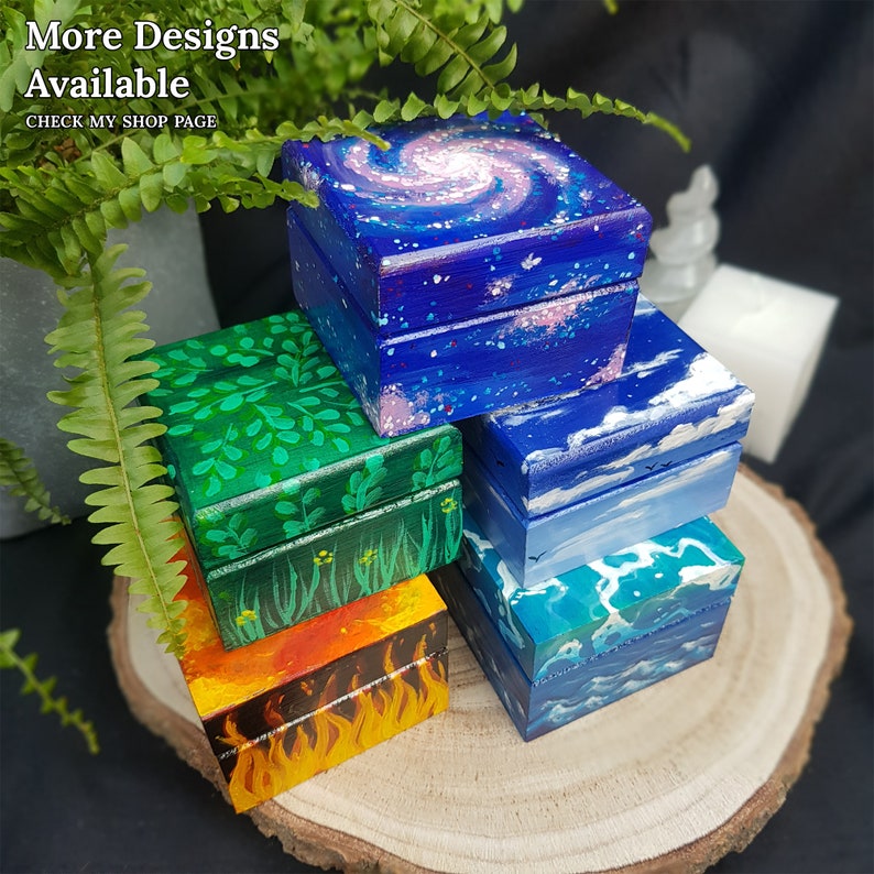 Puede incluir: Una pila de cinco peque&ntilde;as cajas de madera, cada una pintada con un dise&ntilde;o diferente. La caja superior est&aacute; pintada con un dise&ntilde;o de galaxia, la siguiente caja est&aacute; pintada con un dise&ntilde;o de cielo, la siguiente caja est&aacute; pintada con un dise&ntilde;o de oc&eacute;ano, la siguiente caja est&aacute; pintada con un dise&ntilde;o de fuego y la caja inferior est&aacute; pintada con un dise&ntilde;o de bosque.