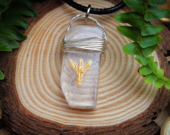 Puede incluir: Un colgante de cristal de cuarzo transparente con un diseño de alambre envuelto en oro y un símbolo de runa dorado. El colgante está en un collar de cordón negro.