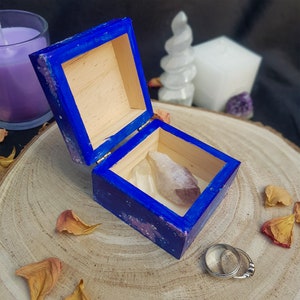 Pu&ograve; includere: Una piccola scatola di legno blu aperta con coperchio a cerniera, contenente un cristallo viola chiaro. La scatola &egrave; decorata con macchie bianche e rosa. Due anelli d'argento sono posizionati accanto alla scatola su una superficie di legno.