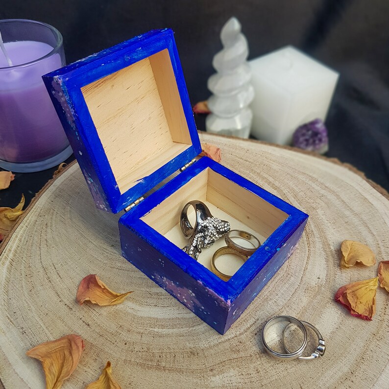 Pu&ograve; includere: Una piccola scatola di legno blu aperta con anelli all'interno, posta su una superficie di legno. La scatola &egrave; dipinta con un motivo maculato blu e viola. Sono visibili diversi anelli, tra cui un grande anello d'argento ornato e due fasce d'oro.
