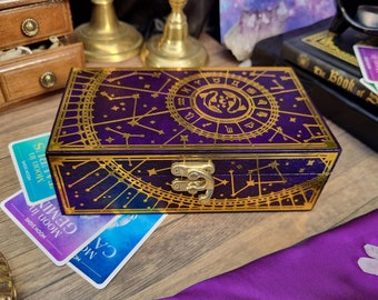 Puede incluir: Una caja rectangular de madera con un acabado morado oscuro y diseños celestiales dorados. La caja tiene una tapa con bisagras y un cierre dorado. Se ven cartas de tarot con el texto "Moon in Gemini" y "Moon in Taurus".