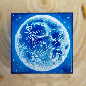 Puede incluir: Una peque&ntilde;a pintura sobre lienzo azul con una luna blanca y estrellas. La luna est&aacute; llena y tiene un dise&ntilde;o blanco detallado en su superficie.