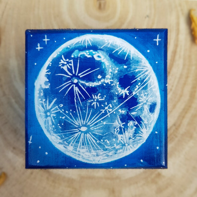 Puede incluir: Una peque&ntilde;a pintura sobre lienzo azul con una luna blanca y estrellas. La luna est&aacute; llena y tiene un dise&ntilde;o blanco detallado en su superficie.