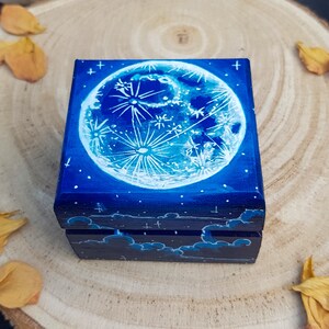 Puede incluir: Una peque&ntilde;a caja de madera con fondo azul y una luna pintada de blanco con estrellas. La luna tiene un dise&ntilde;o detallado de cr&aacute;teres y un dise&ntilde;o en forma de nube en la parte inferior de la caja.