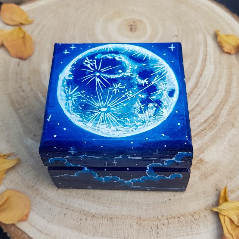 Puede incluir: Una peque&ntilde;a caja de madera con fondo azul y una luna pintada de blanco con estrellas. La luna tiene un dise&ntilde;o detallado de cr&aacute;teres y un dise&ntilde;o en forma de nube en la parte inferior de la caja.
