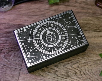 Puede incluir: Una caja de madera negra con un diseño zodiacal plateado en la tapa. El diseño presenta un círculo con los signos del zodíaco y un león en el centro.