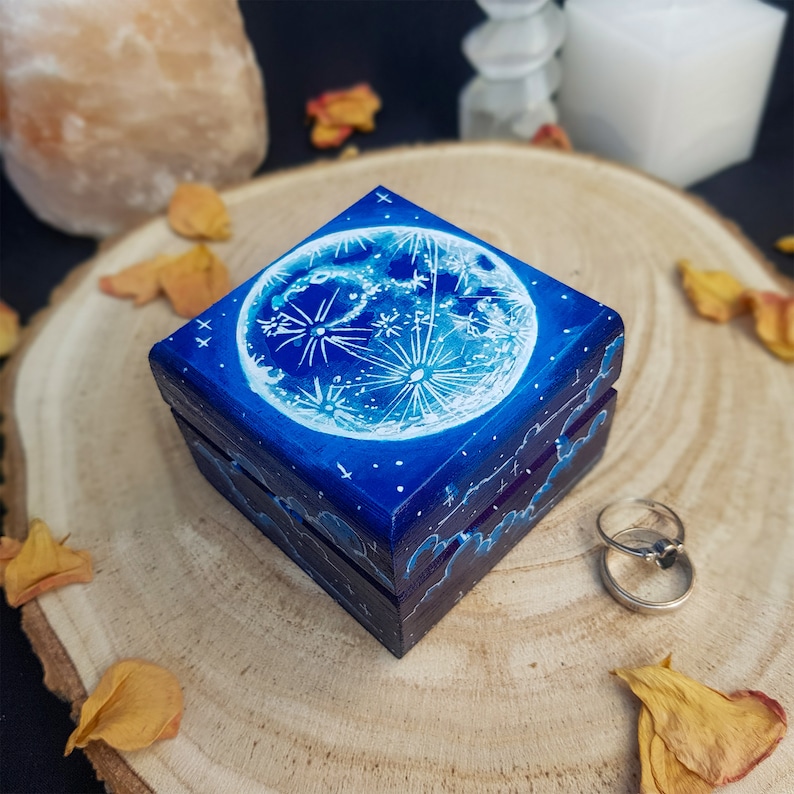 Puede incluir: Una peque&ntilde;a caja de madera con fondo azul y una luna blanca con estrellas pintadas en la tapa. La caja est&aacute; cerrada y se encuentra sobre una superficie de madera.