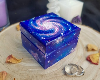 Puede incluir: Una pequeña caja de madera cuadrada pintada con un diseño de galaxia arremolinada en tonos azules, morados y blancos. La caja está abierta, revelando un anillo de plata en el interior. La caja está sobre una superficie de madera.