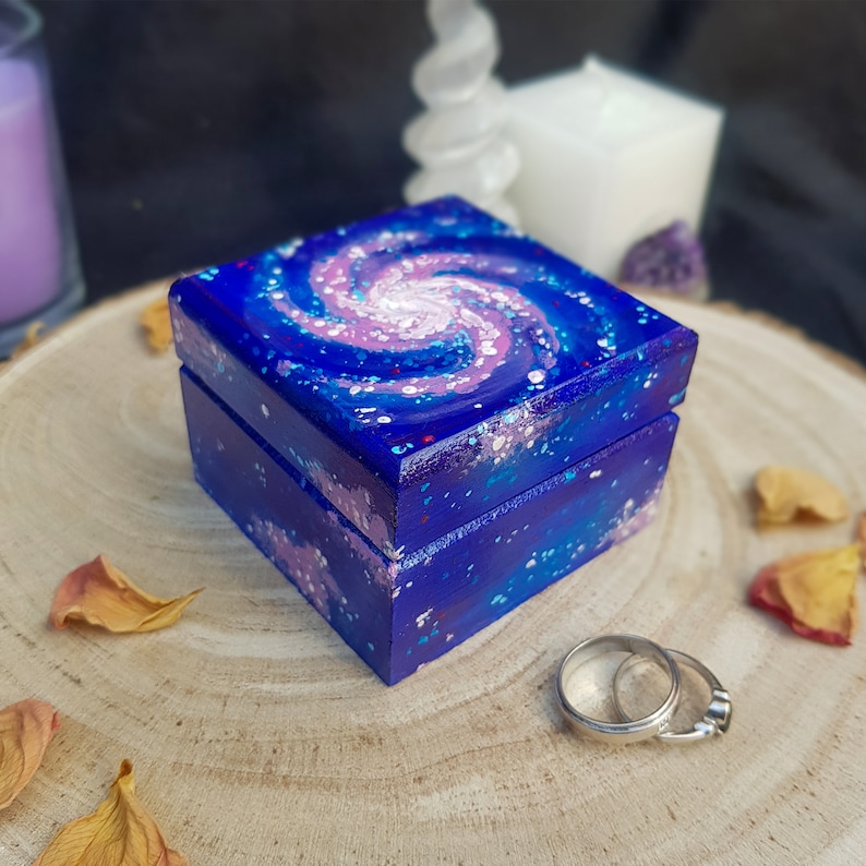 Pu&ograve; includere: Una piccola scatola quadrata di legno dipinta con un motivo a spirale di galassia nei toni del blu, viola e bianco. La scatola &egrave; aperta, rivelando un anello d'argento all'interno. La scatola &egrave; appoggiata su una superficie di legno.