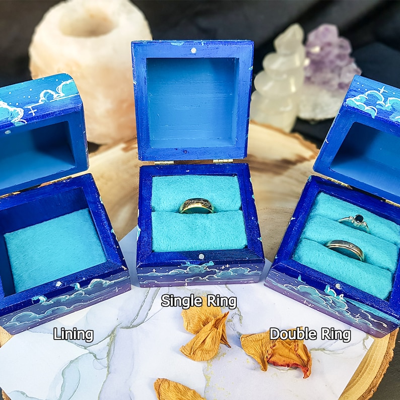 Pu&ograve; includere: Tre scatole portanelli in legno blu con un design a tema cielo. Le scatole sono aperte, rivelando un interno turchese. Una scatola contiene un singolo anello d'oro, un'altra due anelli e la terza &egrave; vuota. Il testo "Single Ring", "Double Ring" e "Lining" &egrave; visibile.