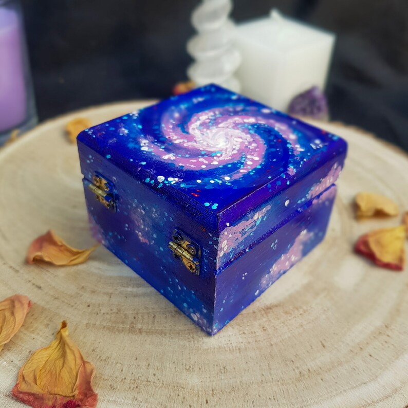 Pu&ograve; includere: Una scatola quadrata di legno dipinta con un design a spirale di galassia nei toni del blu, viola e bianco. La scatola ha cerniere e una chiusura color ottone. La scatola &egrave; appoggiata su una superficie di legno.