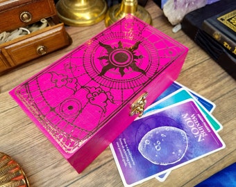 Puede incluir: Una caja de madera rosa con un cierre dorado y un diseño decorativo en la tapa. El diseño presenta un sol con rayos y nubes. La caja está colocada sobre una pila de cartas con un diseño de acuarela púrpura y azul. El texto en la tarjeta dice "Fases lunares Luna creciente Ahora es un buen momento para concentrarse en lograr sus propios objetivos personales."