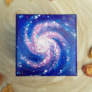 Pu&ograve; includere: Arte su tela quadrata con una vivace pittura di galassia. L'opera d'arte mostra una galassia vorticosa in tonalit&agrave; rosa e bianca su uno sfondo blu scuro, con macchie blu e bianche sparse. Il dipinto evoca un senso di meraviglia cosmica.