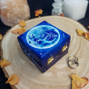 Puede incluir: Una peque&ntilde;a caja de madera con fondo azul y una luna blanca con estrellas pintadas en la tapa. La caja tiene bisagras doradas y est&aacute; cerrada.