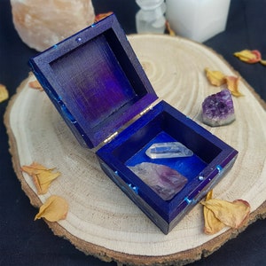 Puede incluir: Una peque&ntilde;a caja de madera con un dise&ntilde;o de galaxia azul y morado. La caja est&aacute; abierta y contiene tres cristales: un cristal de cuarzo transparente, un cristal de amatista morado y un cristal de amatista morado m&aacute;s grande.