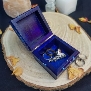 Puede incluir: Una peque&ntilde;a caja de madera azul oscuro con una tapa con bisagras. La caja est&aacute; abierta y contiene dos anillos de plata y un anillo de plata con un dise&ntilde;o decorativo. La caja est&aacute; sentada sobre una superficie de madera marr&oacute;n claro.