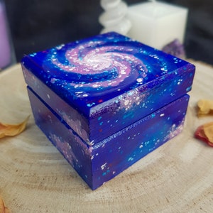 Pu&ograve; includere: Una piccola scatola quadrata di legno dipinta con un vivace design a galassia. La scatola presenta un colore di base blu scuro con un motivo a galassia vorticoso nei toni del rosa, bianco e blu sul coperchio. Le macchie bianche e rosse aggiungono un effetto stellato.