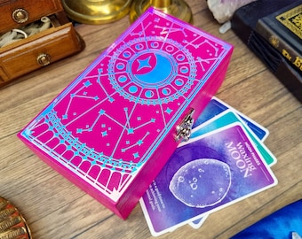 Puede incluir: Una caja de madera rosa con un diseño azul y plateado de una luna creciente y estrellas. La caja está abierta y muestra una baraja de cartas con un diseño morado y azul. Las cartas tienen el texto "Moon Phases Waxing Moon".