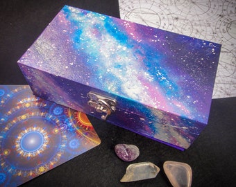Può includere: Una scatola di legno con un design a tema galattico. La scatola è dipinta in tonalità di viola, blu e rosa, e ha una chiusura argentata. La scatola è appoggiata su una superficie nera.