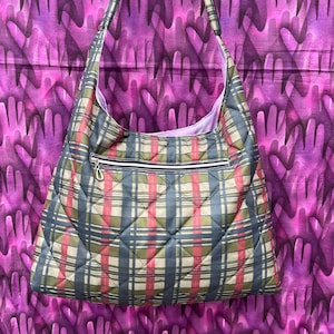 Puede incluir: Un bolso hobo acolchado con un patrón de cuadros en tonos verde oliva, rojo y negro. El bolso cuenta con una sola correa para el hombro, una abertura superior y un bolsillo horizontal con cremallera en el frente con un tirador plateado y forro interior morado claro.