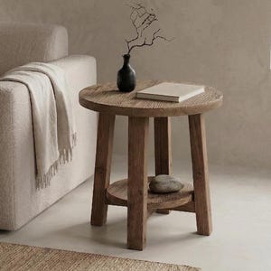 May include: A small, round wooden side table with a lower shelf. A small black vase with a twig and a book sit on top. A stone rests on the shelf. The table is made of light brown wood.