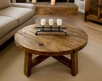 May include: A round wooden coffee table with a black metal candle holder and four white candles. The table has a rustic, distressed finish and sits on a light brown rug in front of a fireplace.