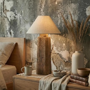 Può includere: Una lampada da tavolo rustica in legno con paralume beige. La lampada è su un comodino in legno con un runner in lino beige. Sullo sfondo, un letto con un piumino in lino beige. Un vaso con piante secche e una piccola ciotola sono anche sul tavolo.