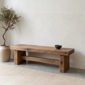 Könnte beinhalten: Eine rustikale Holzbank mit natürlichem Finish. Die Bank wird von dicken, rechteckigen Beinen und einem horizontalen Balken getragen. Eine kleine, dunkle Schale steht auf der Bank. Ein beiger Teppich und ein Topf-Olivenbaum sind ebenfalls im Bild.