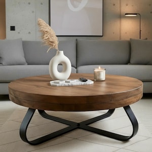 May include: A round wooden coffee table with a dark finish and black metal base. A white vase with a circular opening, a marble tray, a candle, and a remote are on the table. A gray sofa and a rug are in the background.