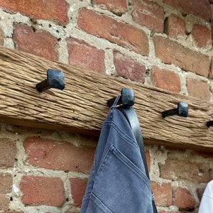 May include: A rustic wooden coat rack with five black metal hooks. The rack is mounted on a brick wall and has a weathered, distressed finish. A blue denim jacket and a white polo shirt are hanging on the hooks.