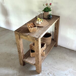 K&ouml;nnte beinhalten: Ein rustikaler Holz-Konsolentisch mit zwei Ablagen. Der Tisch ist mit einer Glasglocke, einem kleinen Teller mit Essen, einer Topfpflanze und mehreren geflochtenen K&ouml;rben dekoriert.