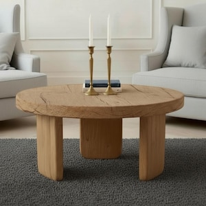 Op de afbeelding: Een ronde, lichtbruine houten salontafel met een getextureerd oppervlak en vier stevige poten. Twee gouden kandelaars met witte kaarsen staan erop, samen met een kleine stapel boeken. De tafel staat in een woonkamer.