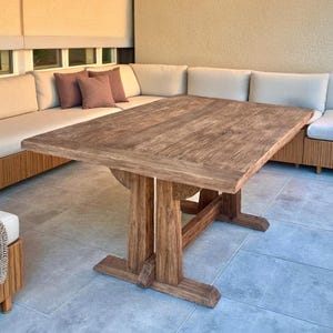 May include: A large, rectangular wooden table with a distressed finish. The table has a sturdy base and is surrounded by a sectional sofa with neutral-colored cushions. The setting is an outdoor patio with tile flooring.