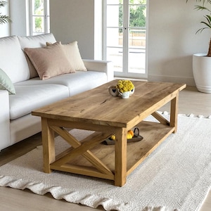 Könnte beinhalten: Rechteckiger Couchtisch aus hellem Holz mit X-förmigem Gestell und unterer Ablage. Er steht auf einem weißen Teppich in einem Wohnzimmer. Im Hintergrund sind ein weißes Sofa und Kissen zu sehen. Der Tisch ist ca. 120 cm lang.