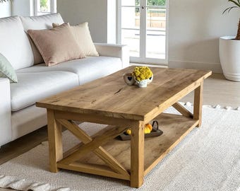 Peut inclure: Table basse rectangulaire en bois clair avec une base en forme de X et une étagère inférieure. Elle est posée sur un tapis blanc dans un salon. Un canapé blanc et des coussins sont visibles en arrière-plan. La table mesure environ 120 cm de long.