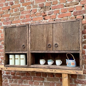 Puede incluir: Un armario de madera con dos puertas y un estante. El armario tiene dos peque&ntilde;os recipientes blancos en el estante y tres tazas peque&ntilde;as y una tetera con un patr&oacute;n floral en el estante inferior. El armario est&aacute; montado en un estante de madera contra una pared de ladrillo.