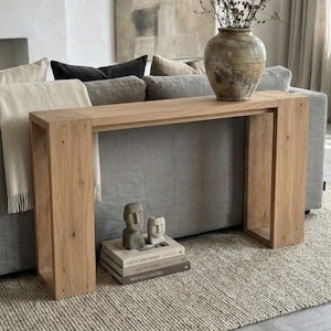 May include: A light oak console table with a rectangular top and open sides. A large, textured vase with dried branches sits on the table. Two small stone sculptures and books are placed below the table. The setting is a living room.