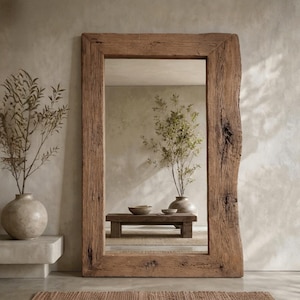 May include: A large rectangular floor mirror featuring a thick, natural-edged wooden frame with visible grain and knots. The mirror reflects a room with a low wooden bench, a ceramic vase, and a plant. A small woven rug sits in front.