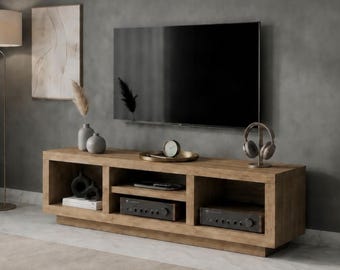 May include: A rectangular wooden media console featuring three open storage compartments. The unit has a natural wood grain finish and sits on a low base. Inside the shelves are electronic components, books, and decorative objects.