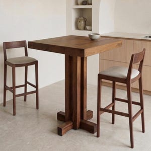 May include: A square, dark wood table with a pedestal base, flanked by two wooden bar stools with light-colored upholstered seats. The stools and table are in a kitchen setting with neutral-toned walls and cabinetry. A small bowl sits on the table.