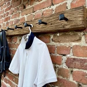 May include: A rustic wooden coat rack with black metal hooks. The rack is mounted on a brick wall and has a white shirt and blue jeans hanging from it.