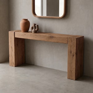 May include: A rectangular console table crafted from light brown wood with a visible grain and textured finish. The table features two thick, block-style legs and a flat top surface. A terracotta-colored vase and a small abstract sculpture sit on top.