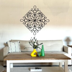 May include: A white sofa with a black metal wall decoration above it. A coffee table with a white base and a wooden top sits in front of the sofa. There are two vases on the table, one yellow and one green. A book is on the lower shelf of the table.