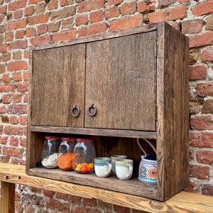 Puede incluir: Un armario de pared de madera r&uacute;stica con dos puertas y un estante. El armario tiene dos tiradores de anillo de metal y est&aacute; pintado de un color marr&oacute;n oscuro. El estante est&aacute; lleno de tarros de vidrio y tazas.