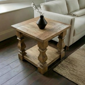 Può includere: Un tavolino quadrato in legno con un ripiano inferiore e gambe tornite decorative. Il tavolo è in legno marrone chiaro e ha un vaso marrone scuro con fiori bianchi. Il tavolo misura circa 61 cm quadrati.