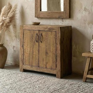 May include: A rustic wooden cabinet with two doors and metal handles. The cabinet is made of weathered wood and has a natural finish. A wooden framed mirror hangs above the cabinet. A vase with dried plants and a small wooden stool are also visible.
