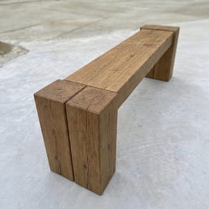 May include: A rectangular bench constructed from thick, solid brown timber beams. The design features a long horizontal seat supported by two sturdy, block-style legs at each end, showcasing a natural wood grain texture and visible end grain patterns.