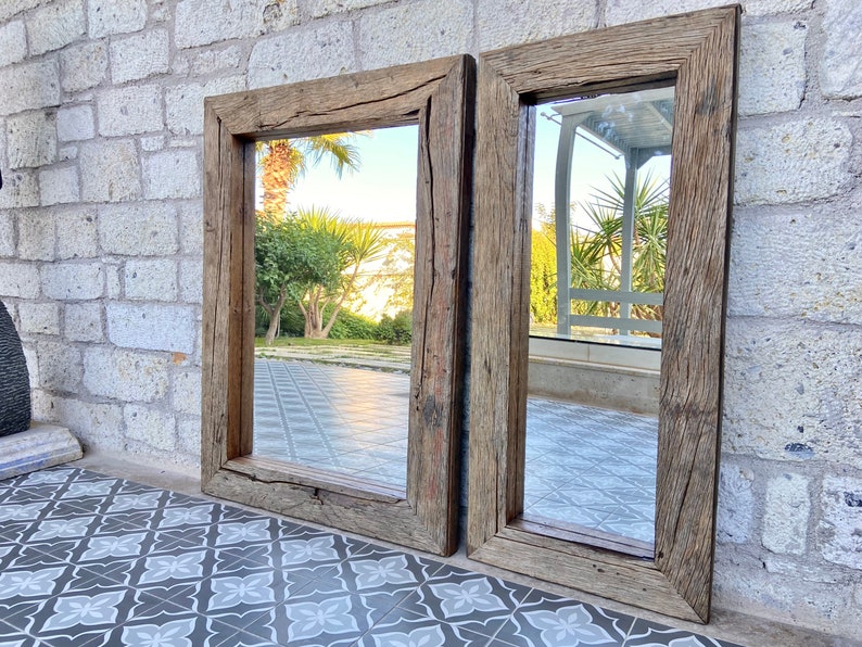Puede incluir: Dos espejos rectangulares con marco de madera r&uacute;stica. Los espejos est&aacute;n colgados en una pared de piedra y reflejan una vista de un patio con plantas y un suelo de baldosas.