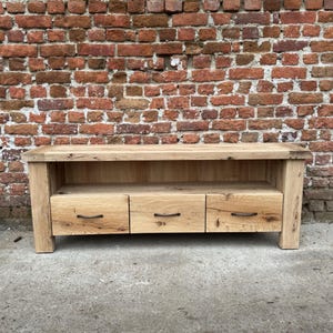 May include: A light-colored wooden media console with three drawers and a shelf. The console has a rectangular shape and is made of natural wood. The drawers have black handles. The background is a brick wall.